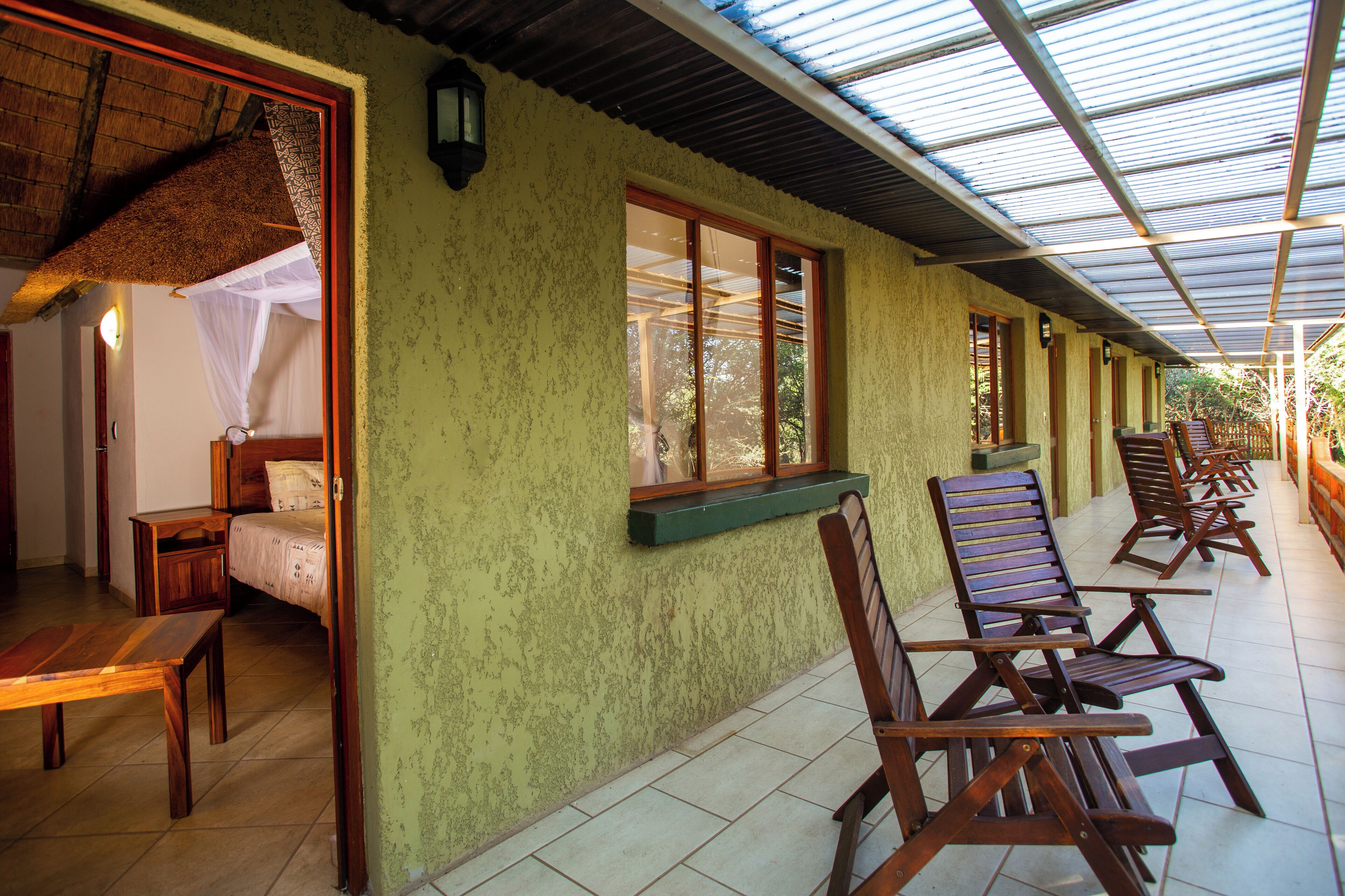 A room with a bed, a table, and a window. There are wooden chairs outside the room.