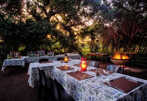 An outdoor dining area with tables set for a meal, surrounded by trees and a fire pit. - Kubu Safari Lodge (Hoedspruit)