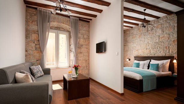 Une pièce avec un mur en pierre, des poutres en bois, un lit recouvert d’une courtepointe bleue, un canapé gris et une petite table sur laquelle est posé un vase de fleurs.