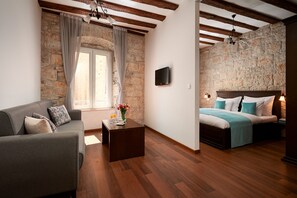 A room with a stone wall, wooden beams, a bed with a blue bedspread, a grey sofa, and a small table with a vase of flowers. - Villa Olea (Split)