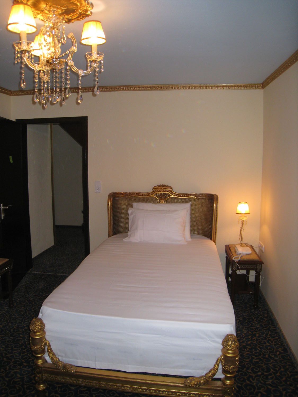 A hotel room with a large bed, a chandelier, a bedside table with a lamp, and a door leading to another room.