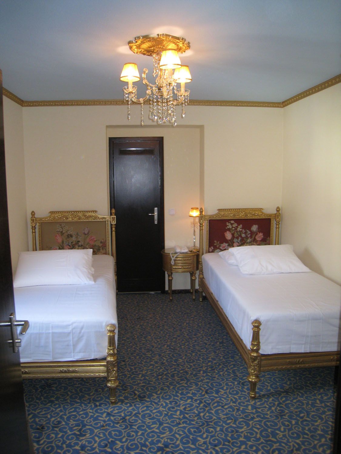 Quarto de hotel com duas camas de solteiro, um candelabro, uma porta e uma pequena mesa de cabeceira com abajur.