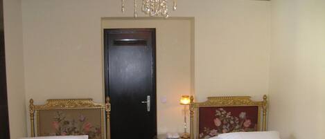 Quarto de hotel com duas camas de solteiro, um candelabro, uma porta e uma pequena mesa de cabeceira com abajur.