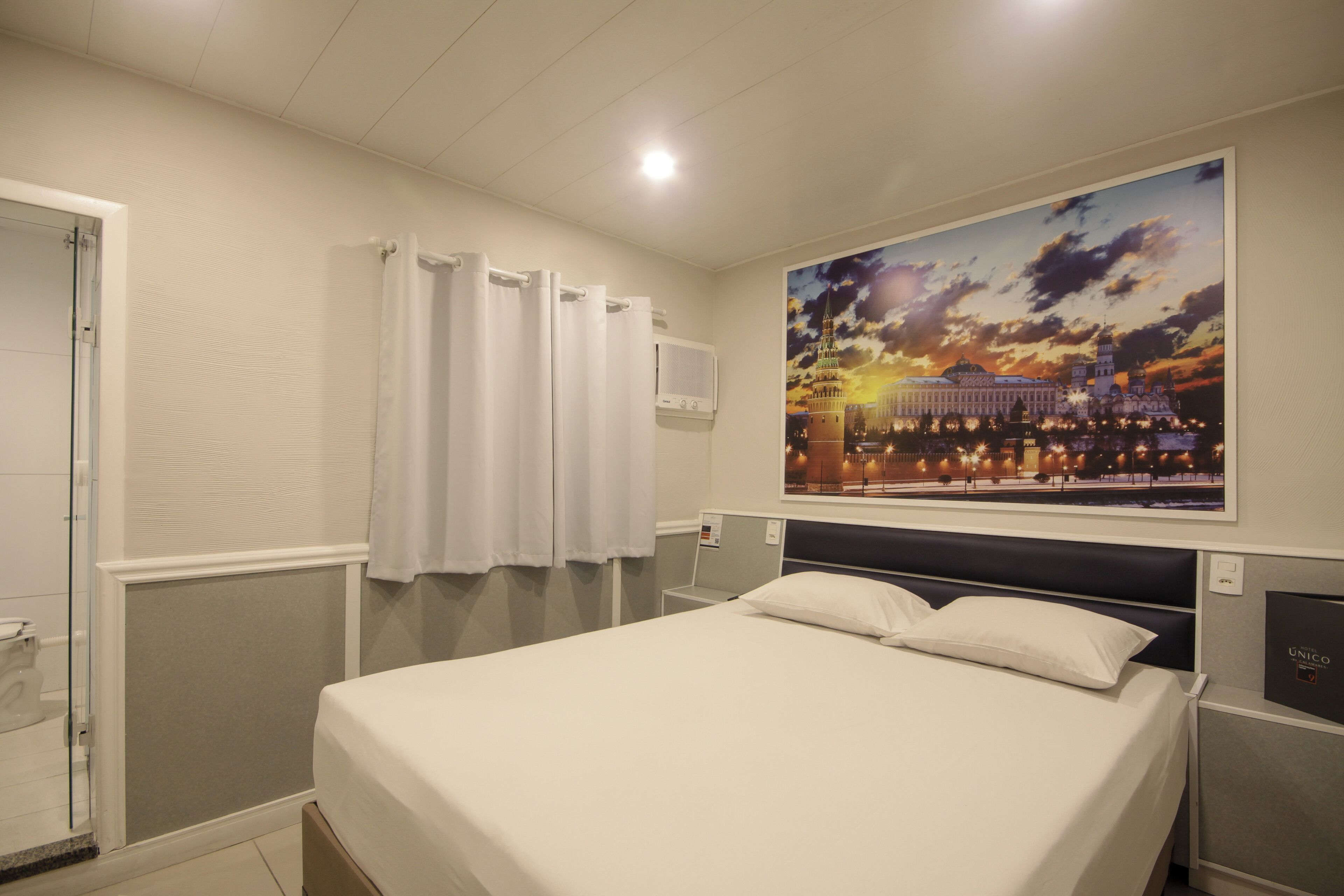A hotel room with a bed, a shower, a window with curtains, and a wall-mounted air conditioner.