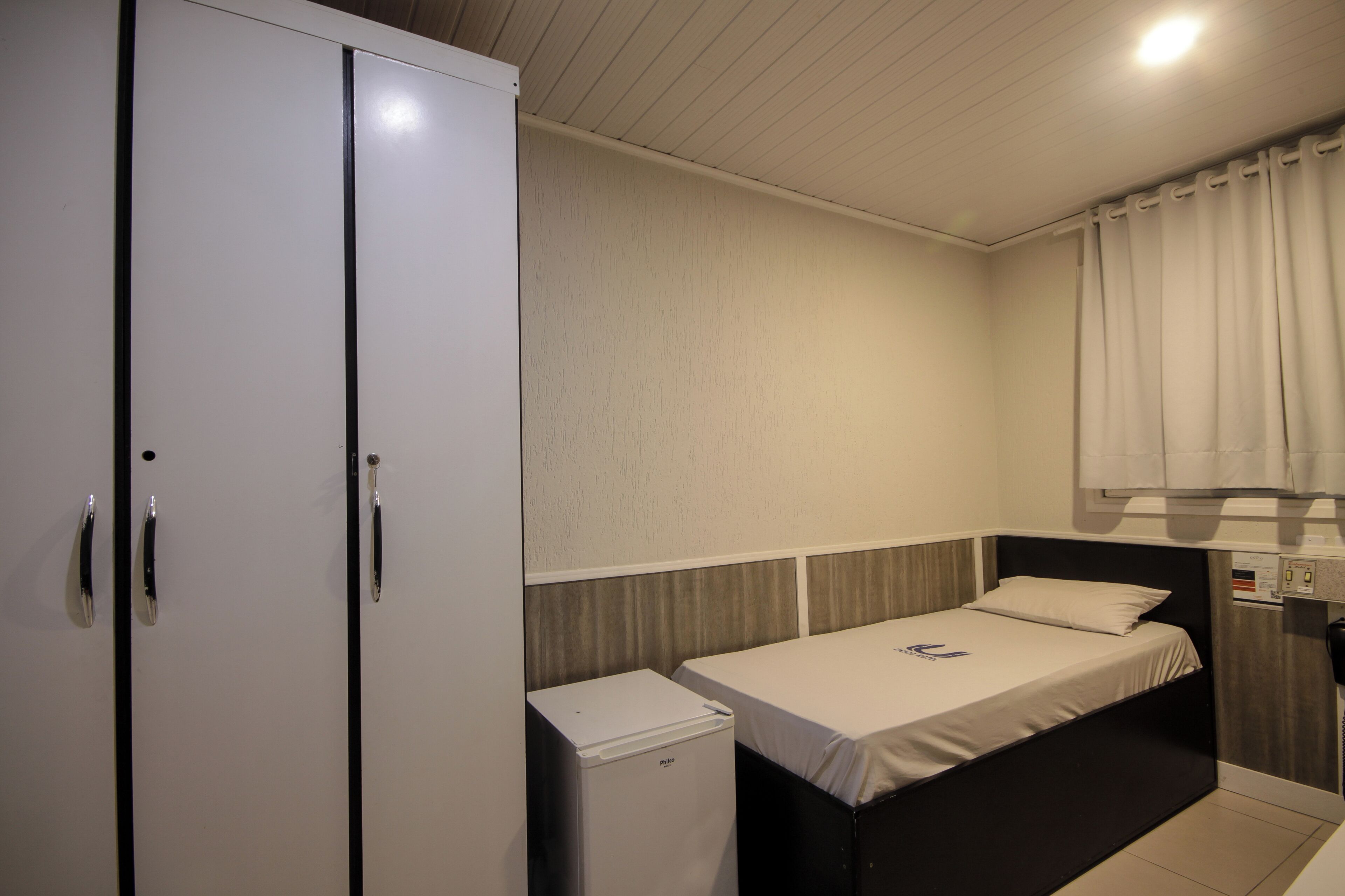Una habitación pequeña y funcional con una cama individual, un armario y un refrigerador compacto.