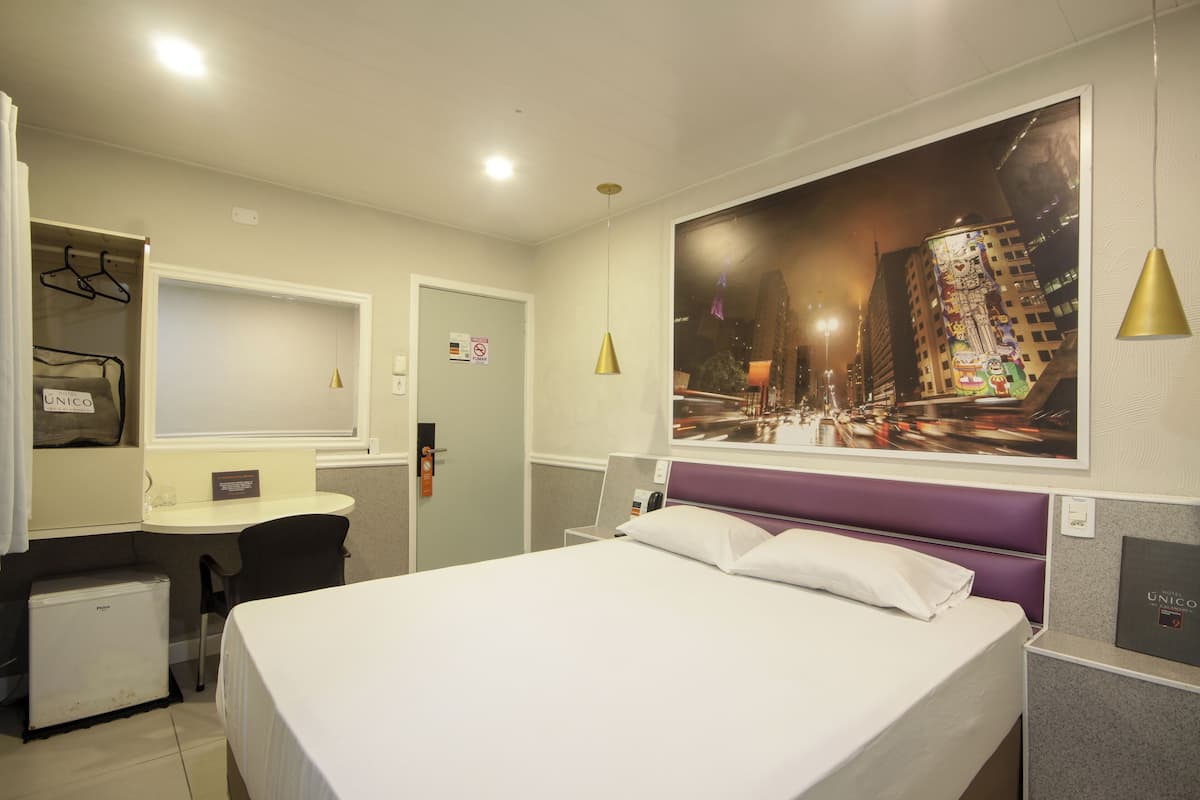 A hotel room with a bed, a desk, a chair, a small fridge, and a large cityscape painting on the wall.