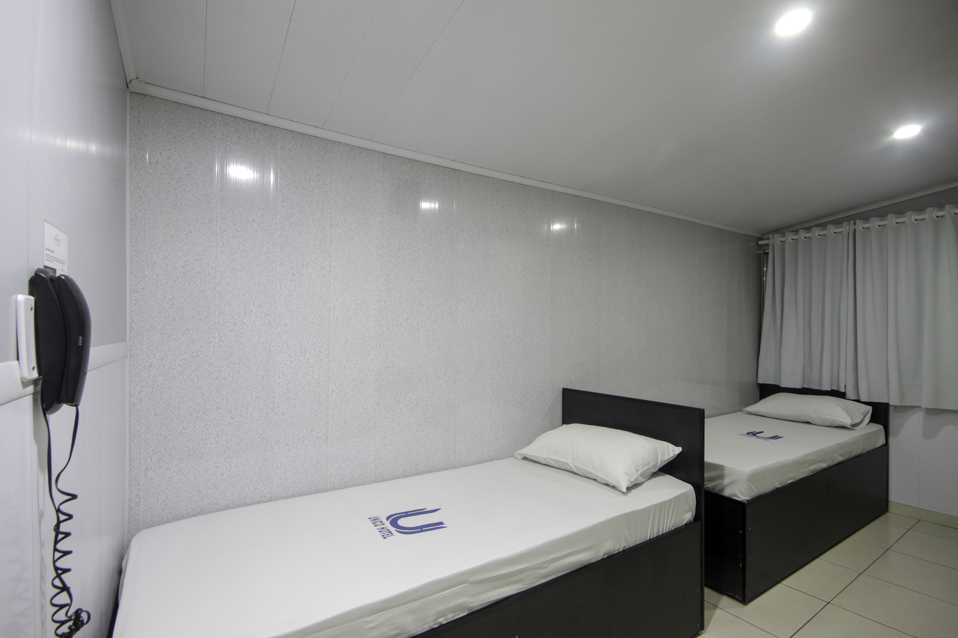 Une chambre avec deux lits simples, un téléphone fixé au mur et un rideau à la fenêtre.