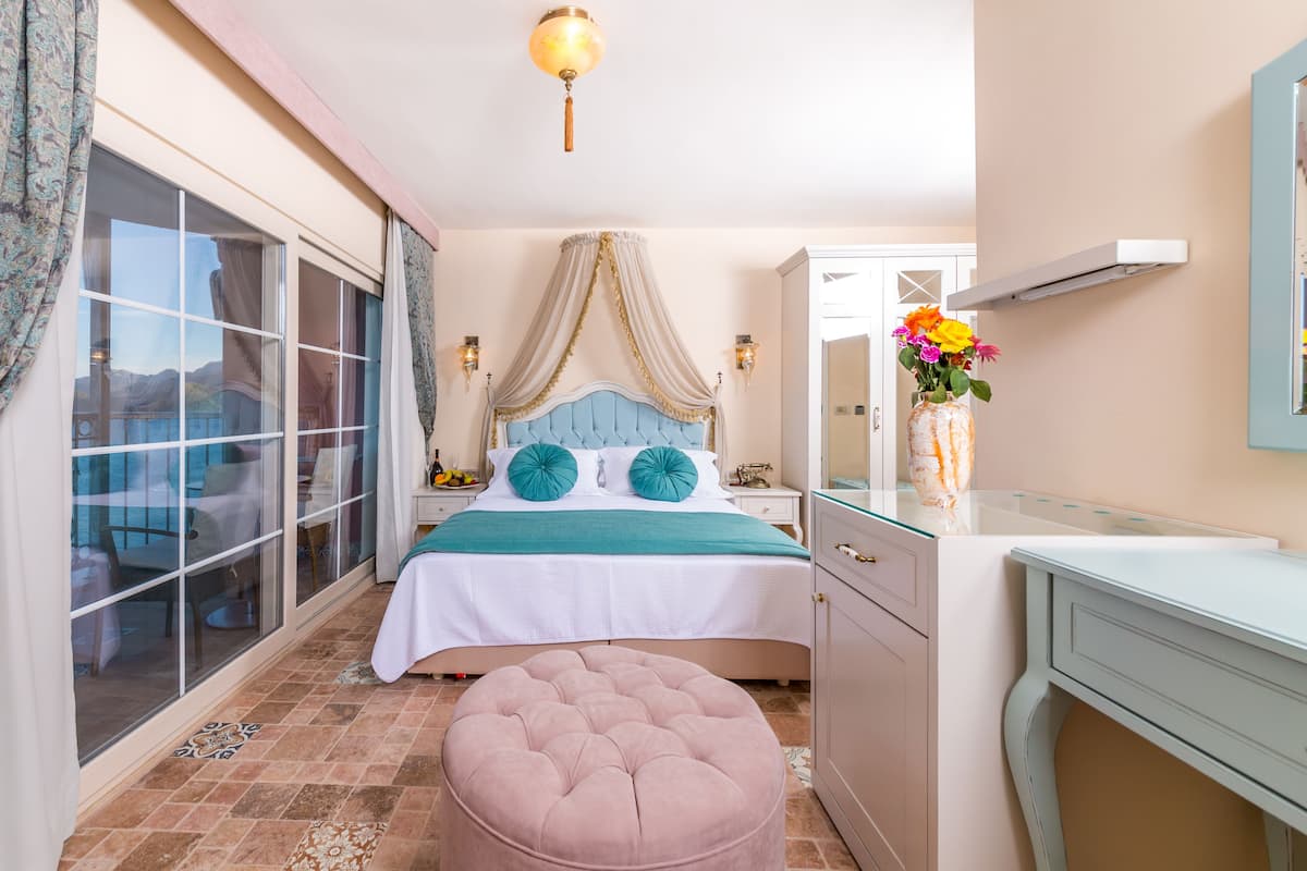 A bedroom with a large window, a bed with teal bedding, a pink ottoman, and a vanity with a mirror.