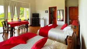 Een hotelkamer met twee bedden, een plafondventilator, een televisie, een eettafel met stoelen, en uitzicht op de omgeving.