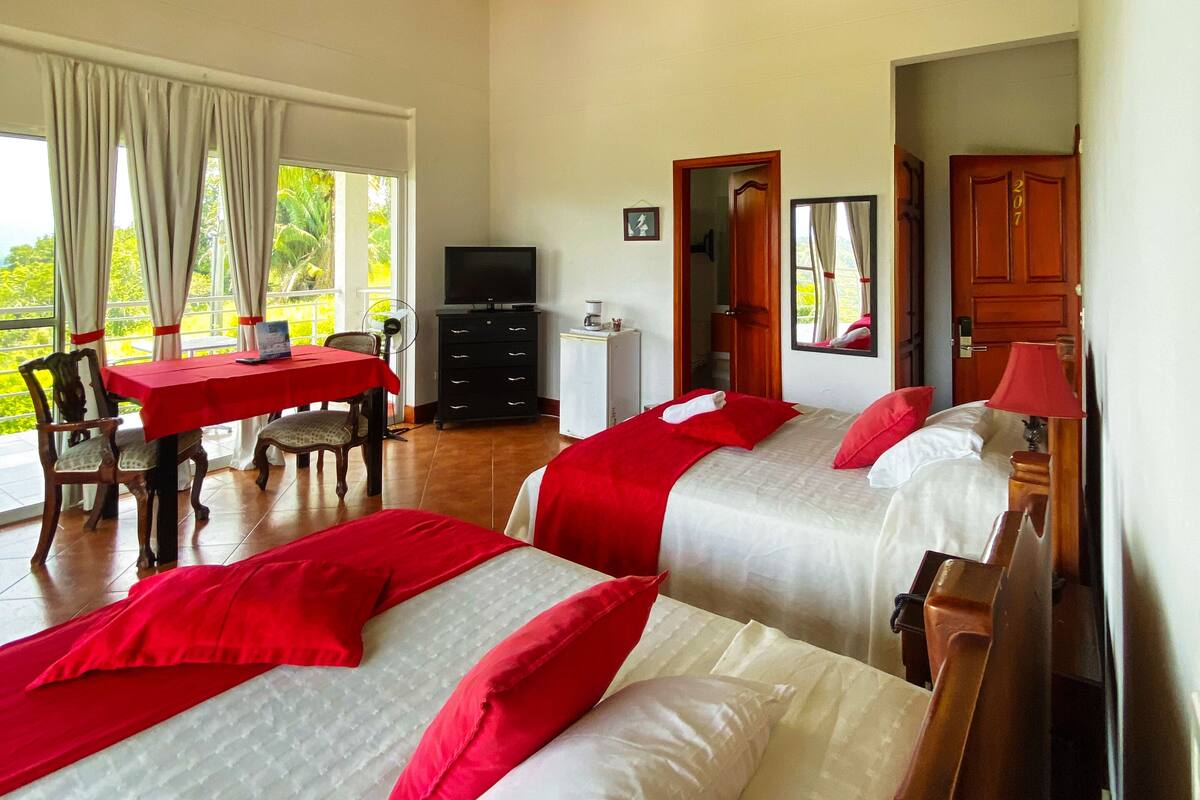 Habitación de hotel con dos camas, ventilador de techo, televisión, mesa de comedor con sillas y vistas al exterior.
