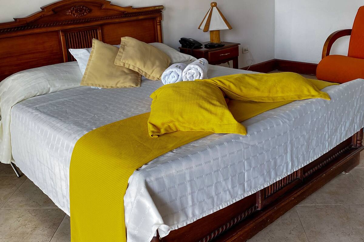 Una cama bien hecha con una colcha blanca y amarilla, un cabecero de madera y una mesita de noche con lámpara y teléfono.
