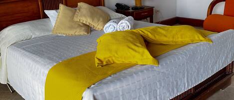 Un lit bien fait, recouvert d’une couvre-lit blanche et jaune, avec une tête de lit en bois et une table de chevet sur laquelle se trouvent une lampe et un téléphone.