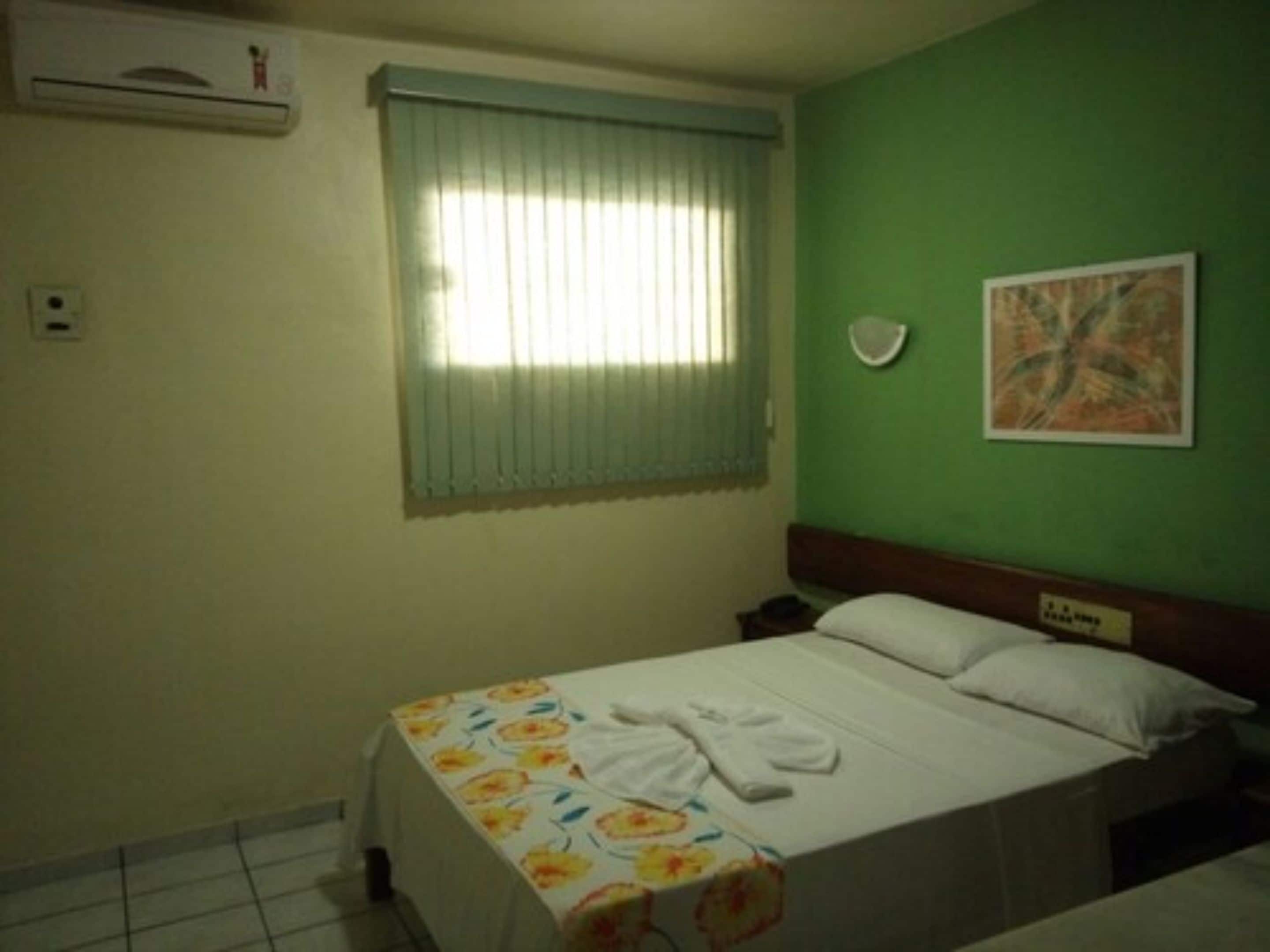 A hotel room with a bed, a window with blinds, a wall-mounted air conditioner, and a painting.