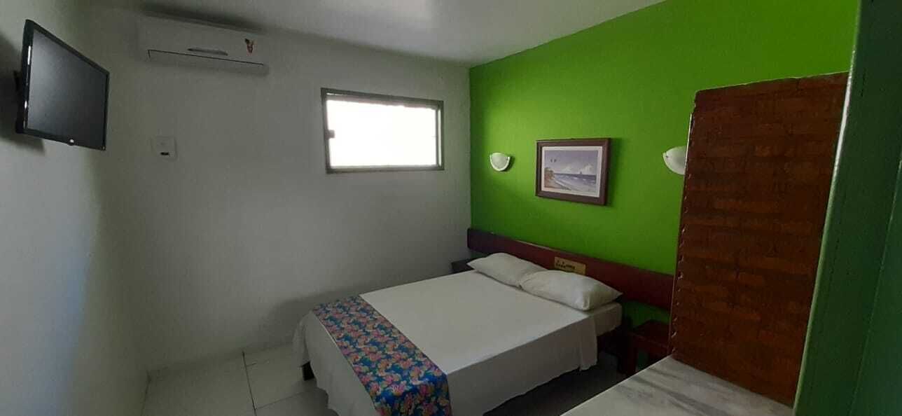 A small, simple room with a bed, a window, a flat-screen TV, and a green wall.