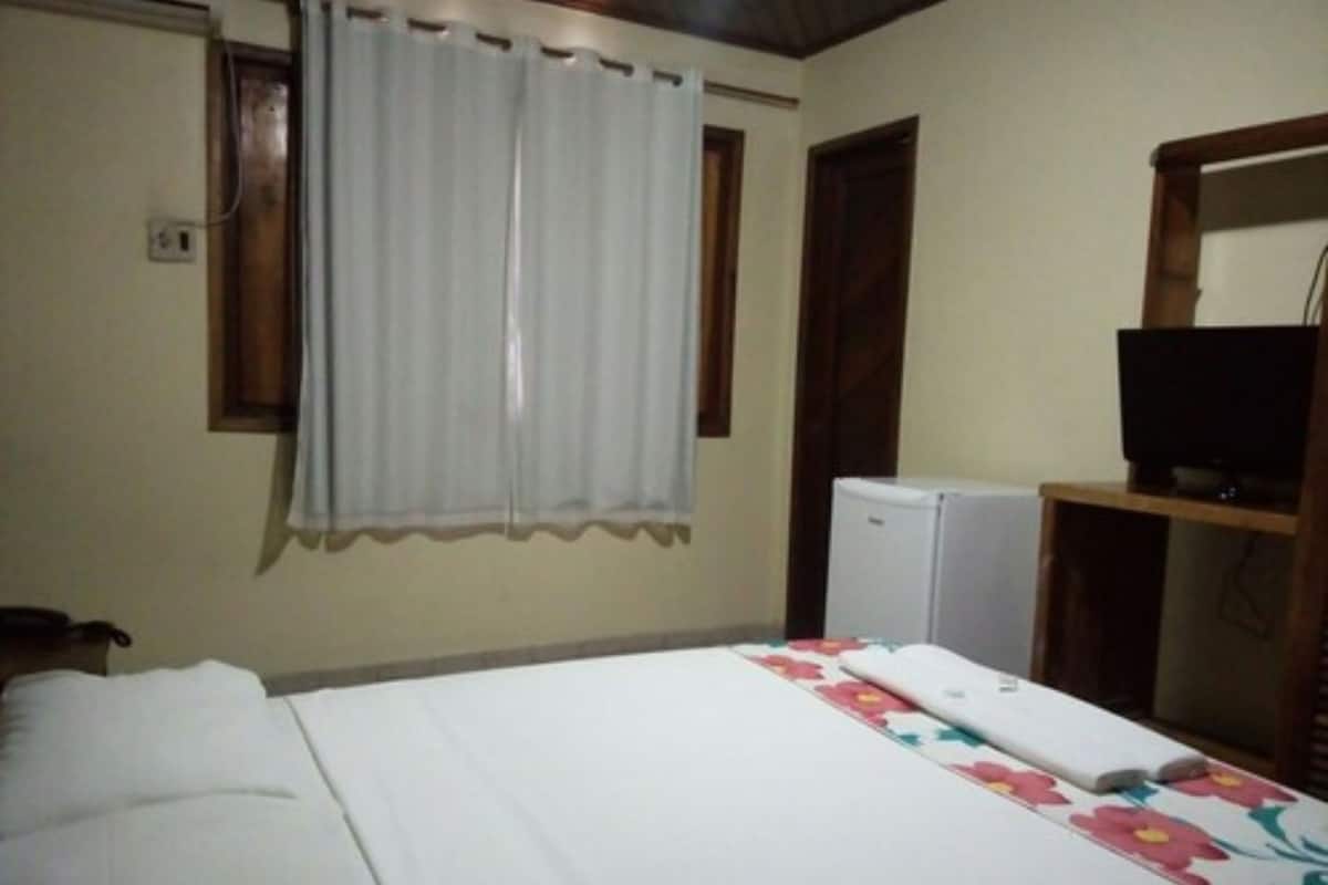 Ein Schlafzimmer mit einem Bett, einem Fernseher auf einem Schrank, einem kleinen Kühlschrank und einem Fenster mit Vorhängen.