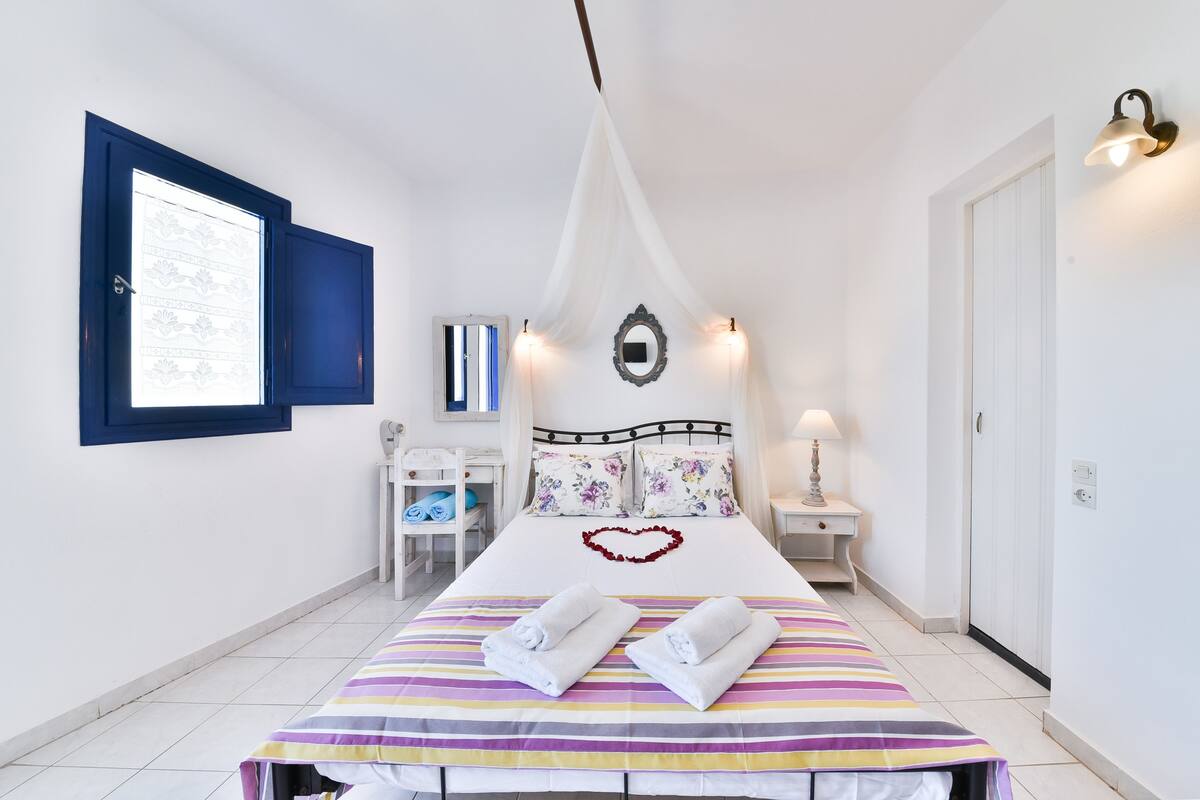 Ein Hotelzimmer mit einem Bett, einem Nachttisch mit Lampe, einem Sessel, einem Fenster mit blauen Rollläden und einem wandmontierten Spiegel.