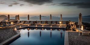 Uma área de piscina tranquila com espreguiçadeiras e guarda-sóis, uma piscina de pedra e vista para o mar ao pôr do sol.