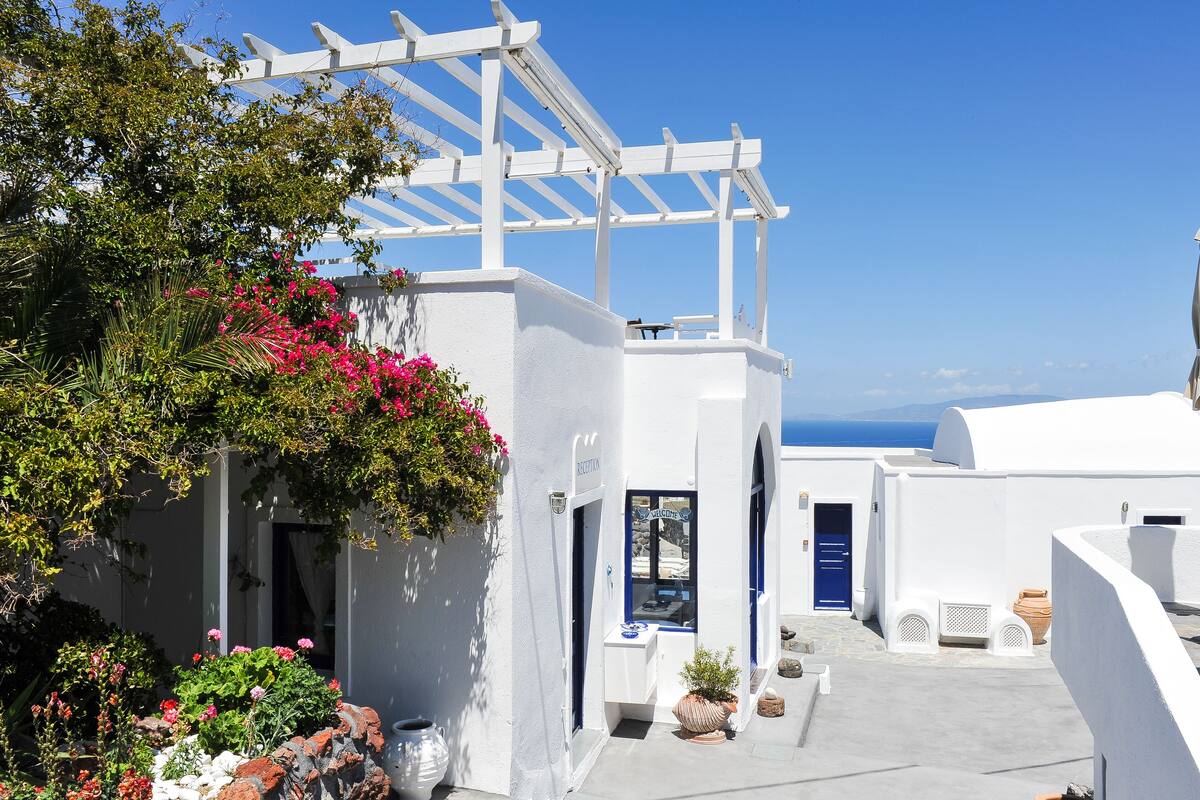 Ein weißes Gebäude mit blauer Tür, einer Pergola und Blick aufs Meer.