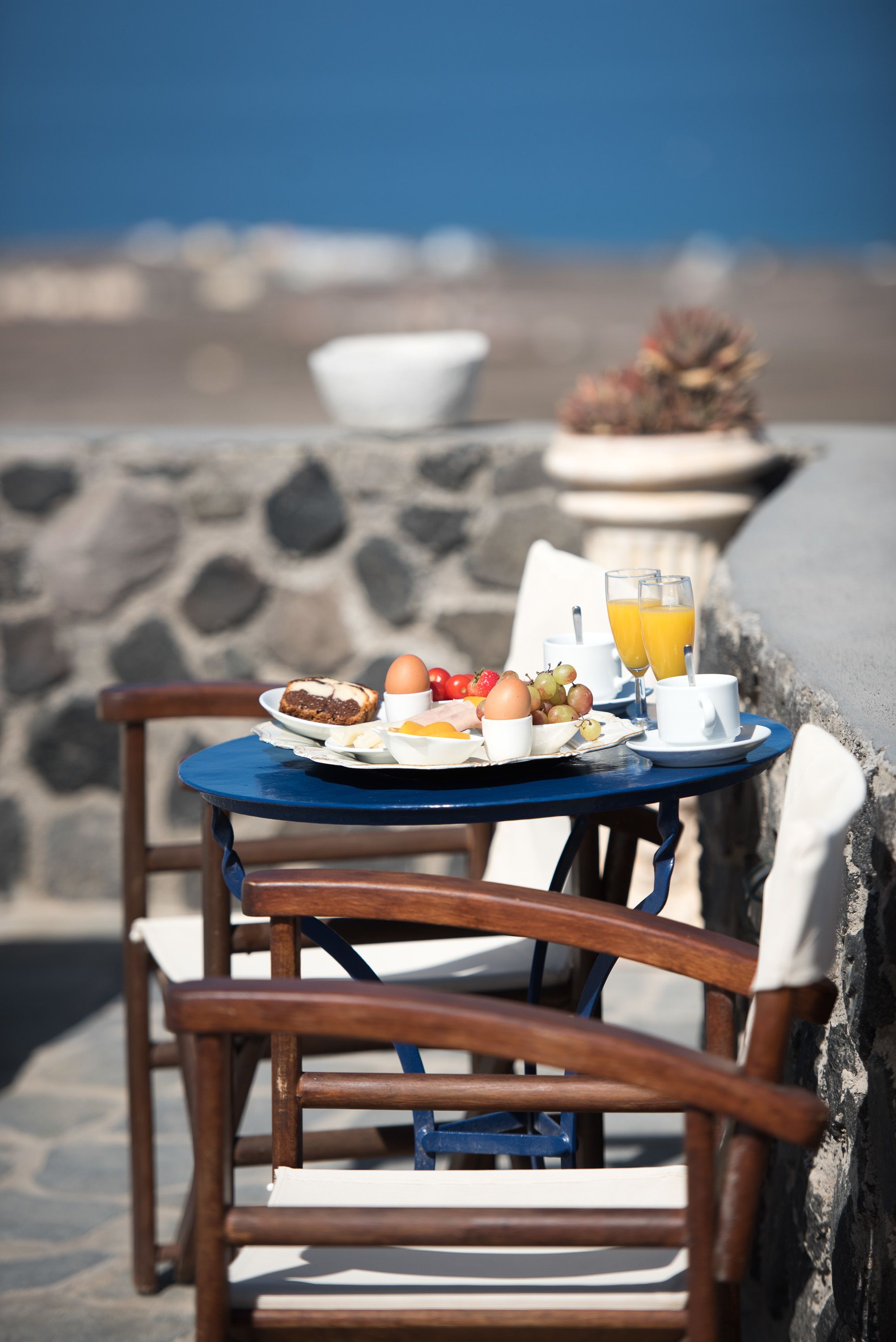 A table with breakfast items and a glass of orange juice, set against a stone wall with a view of the sea.