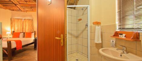 Habitación de hotel con puerta de madera, cama con ropa de cama naranja, zona de ducha con piso de baldosas y lavamanos con espejo.