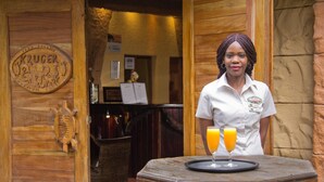La entrada de un hotel con una puerta de madera, un letrero que dice "Kruger Adventure Lodge" y una mesa con dos vasos de jugo de naranja.