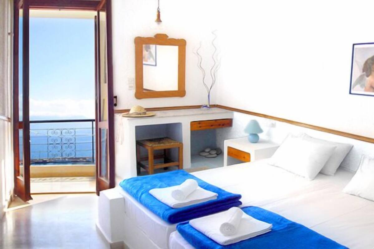 Une chambre d’hôtel avec un lit, un bureau, une chaise, une petite table, une lampe, un tableau au mur et une vue sur la mer.