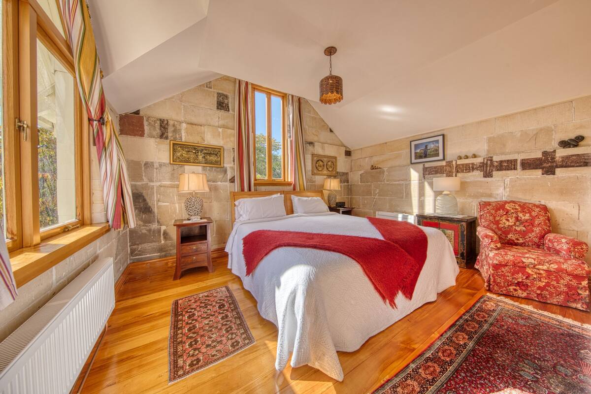 Quarto com uma cama grande, piso de madeira, paredes de pedra, uma janela com cortinas, uma cadeira vermelha e um lustre.