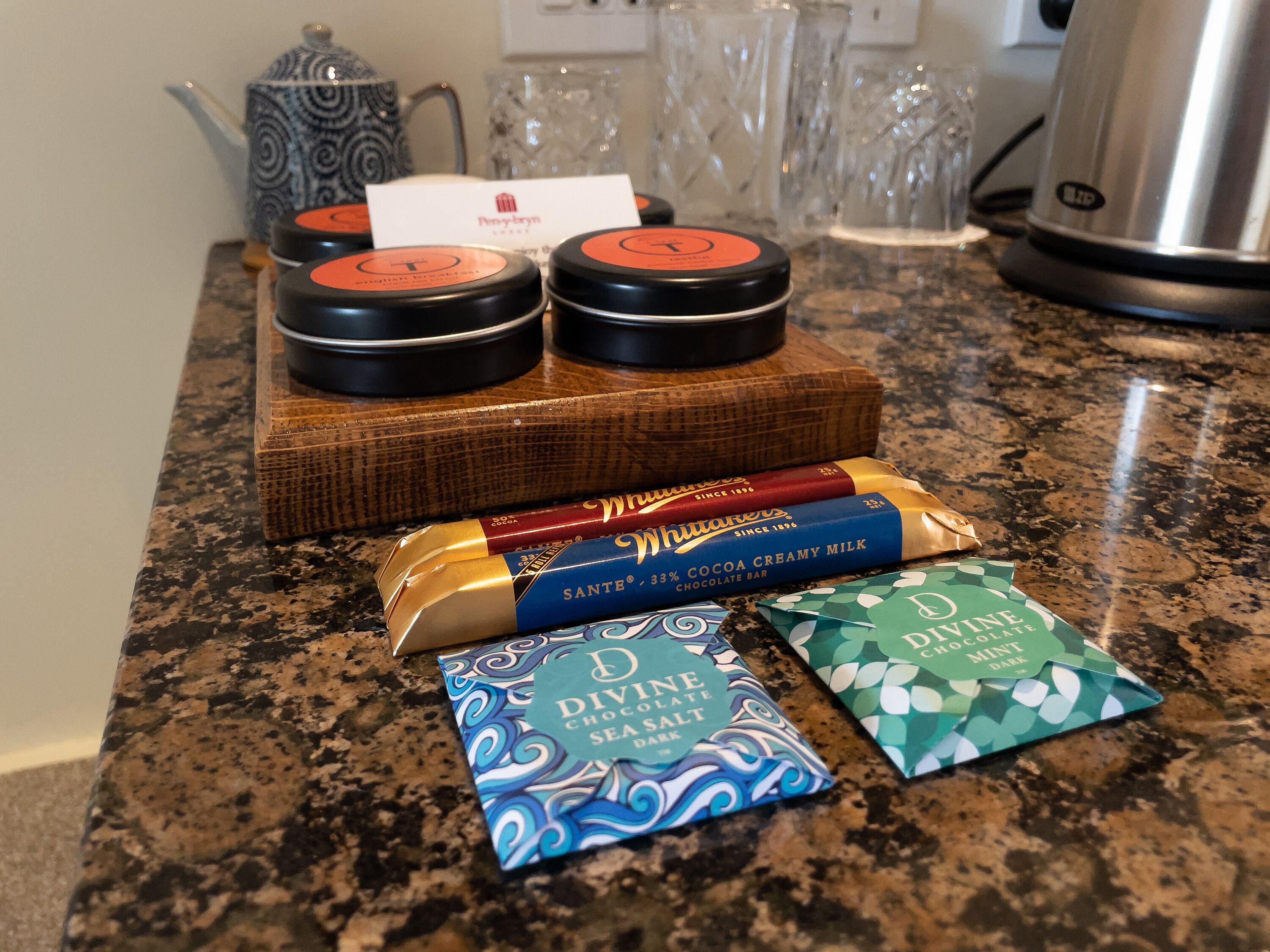 A kitchen counter with a wooden cutting board holding three tins of tea, a bar of chocolate, and two small boxes of chocolate salt.