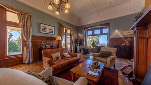 A spacious living room with a red carpet, a wooden coffee table, a sofa, and a chair. There are windows with curtains, a chandelier, and a fireplace. - Pen-y-bryn Lodge (Oamaru)