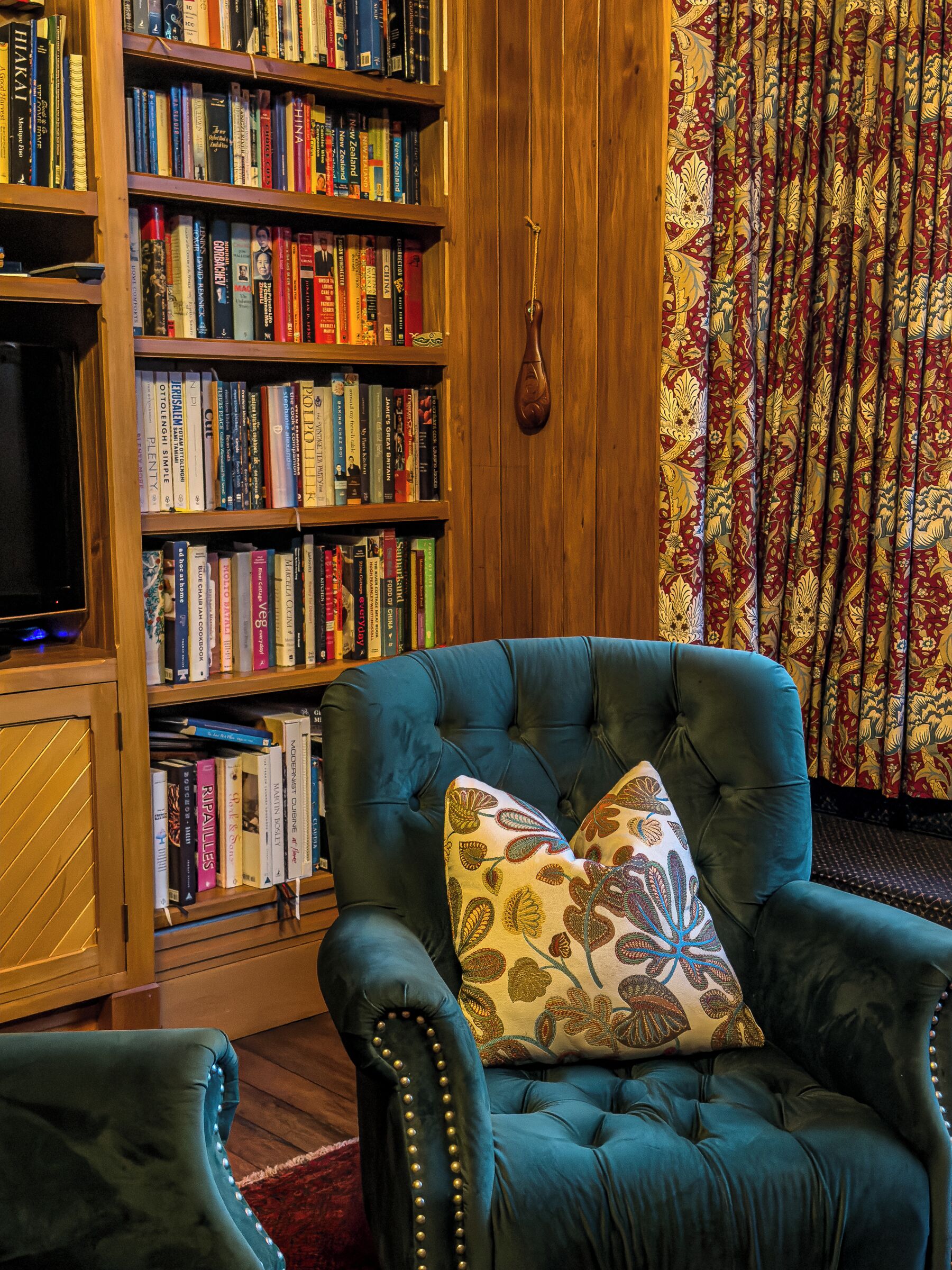 A room with a large bookshelf filled with books, a tufted armchair with a patterned pillow, and a wooden wall.