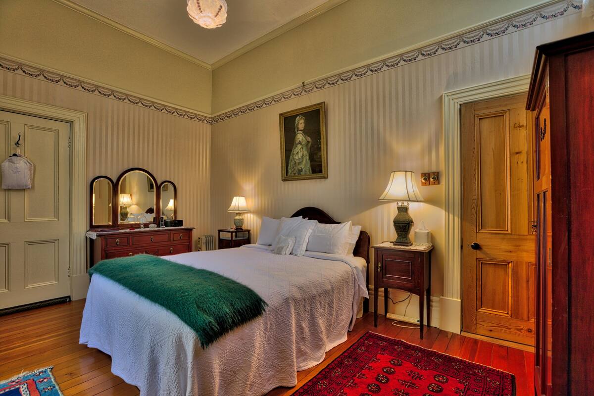 Quarto com uma cama grande, cômoda, duas mesas de cabeceira com abajures, uma porta e um quadro na parede.