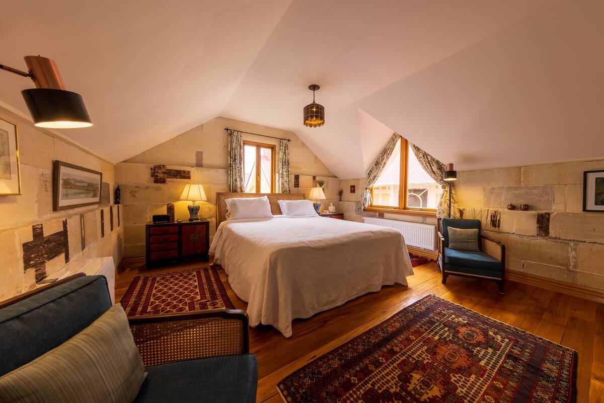 Um quarto de sótão aconchegante com uma cama grande, uma poltrona azul, piso de madeira e teto inclinado.