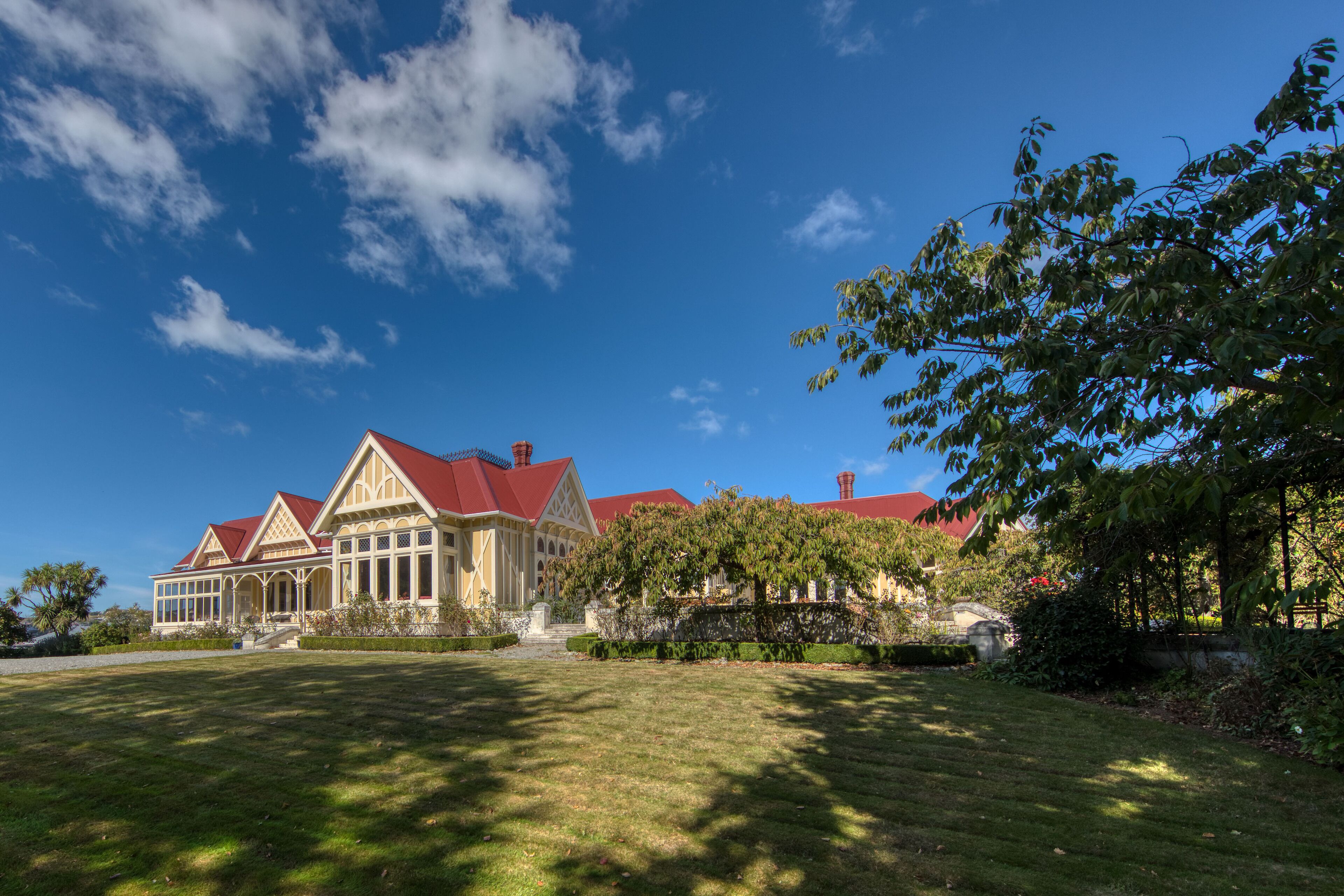 A large, elegant house with a red roof and multiple chimneys, surrounded by well-maintained lawns and trees.