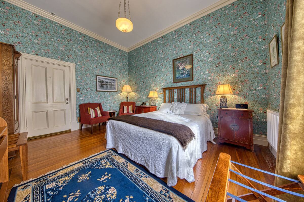 Quarto com papel de parede floral, cama de madeira, duas mesas de cabeceira com luminárias, uma cadeira vermelha e um tapete azul estampado.