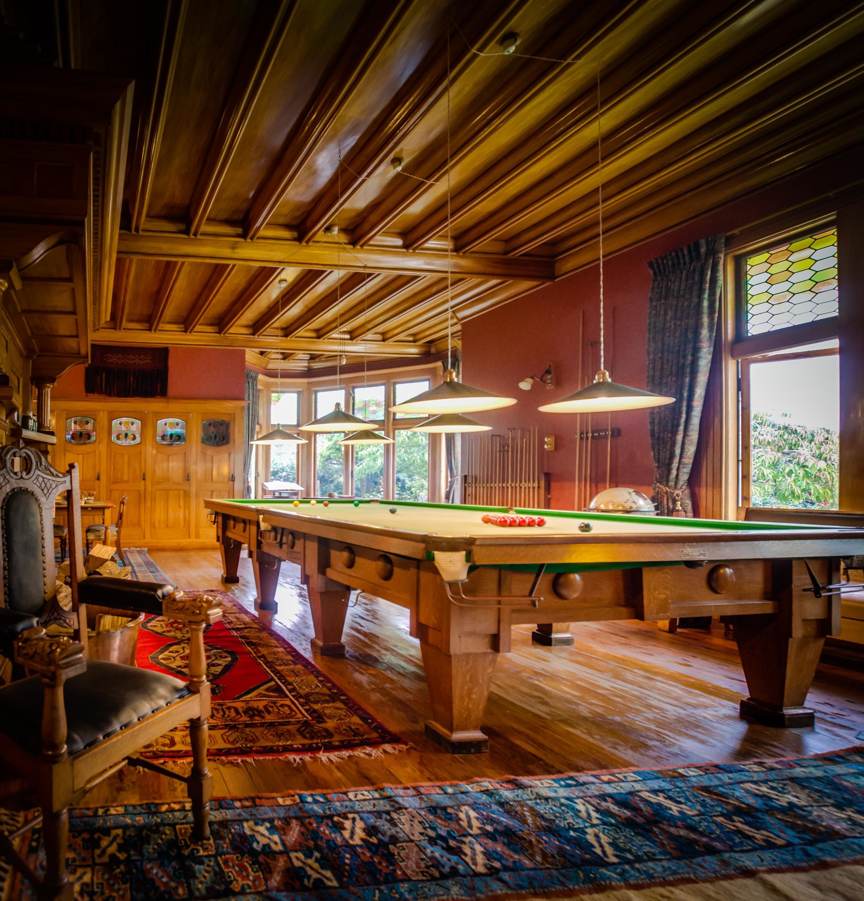 A pool table in a room with wooden beams, a billiards table, and a window with a view of greenery.