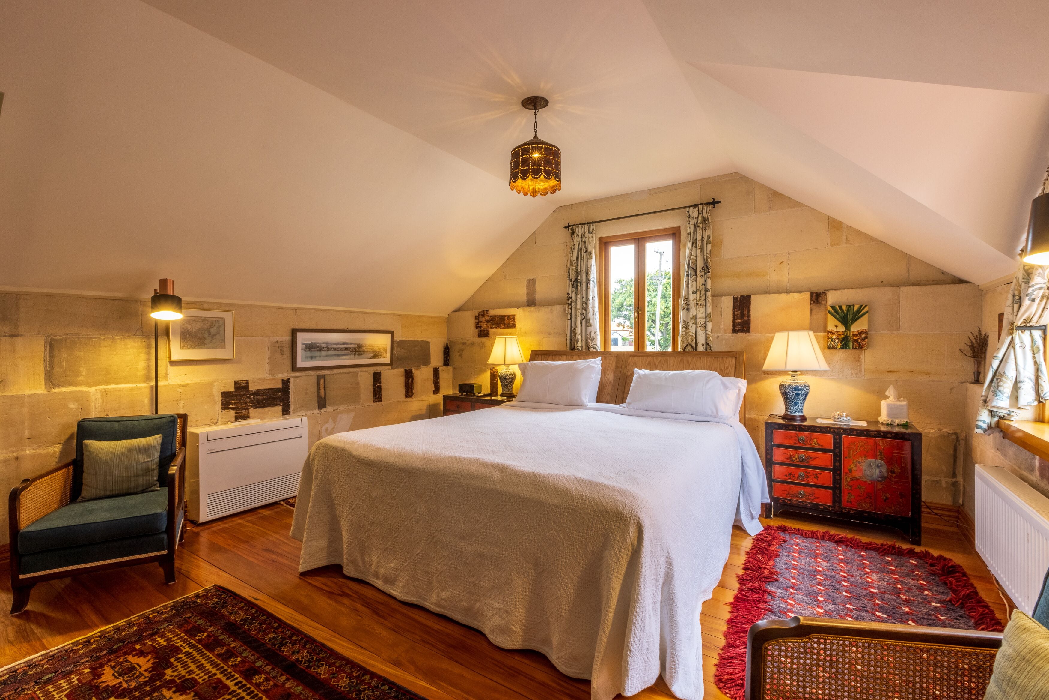 A cozy bedroom with a large bed, a wooden floor, a chair, a nightstand with a lamp, and a window with curtains.