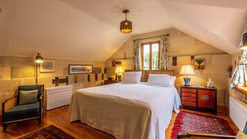 A cozy bedroom with a large bed, a wooden floor, a chair, a nightstand with a lamp, and a window with curtains.