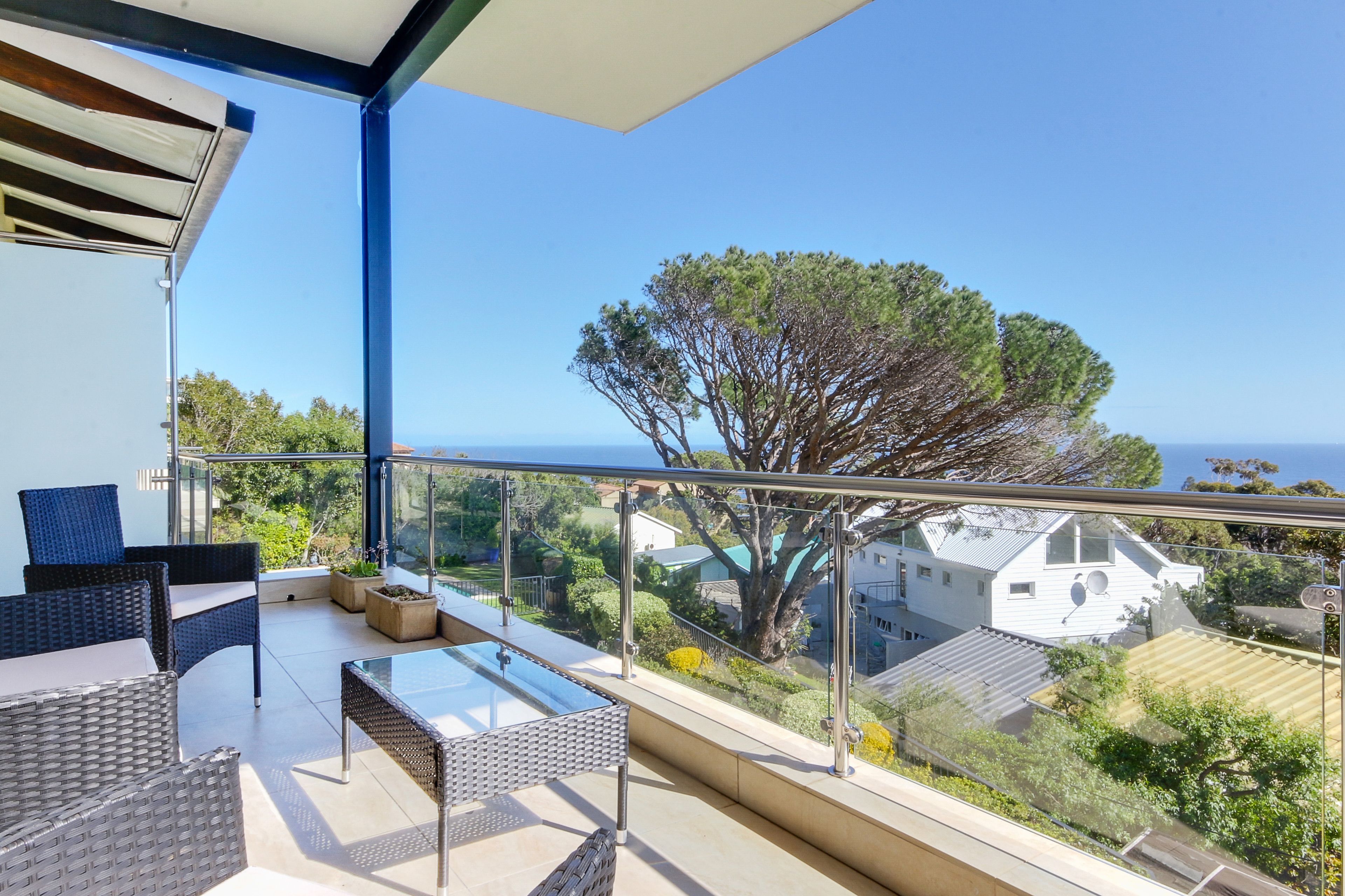 A balcony with wicker furniture, a glass table, and a view of the sea and surrounding houses.