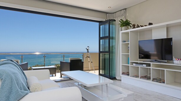 Een moderne woonkamer met een grote televisie, witte meubels en een balkon met uitzicht op de oceaan.
