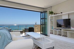 Uma sala de estar moderna com uma televisão grande, móveis brancos e uma varanda com vista para o oceano.