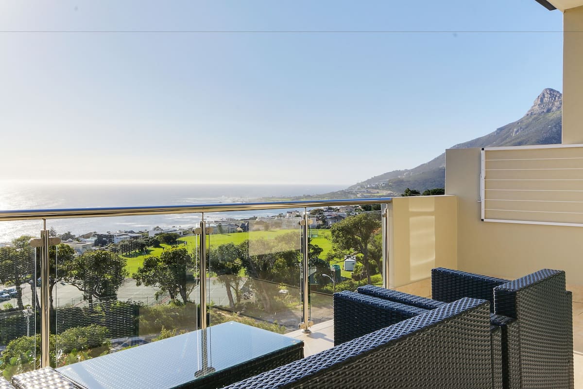 A balcony with a clear view of the ocean and a mountain. There are wicker chairs and a glass table.