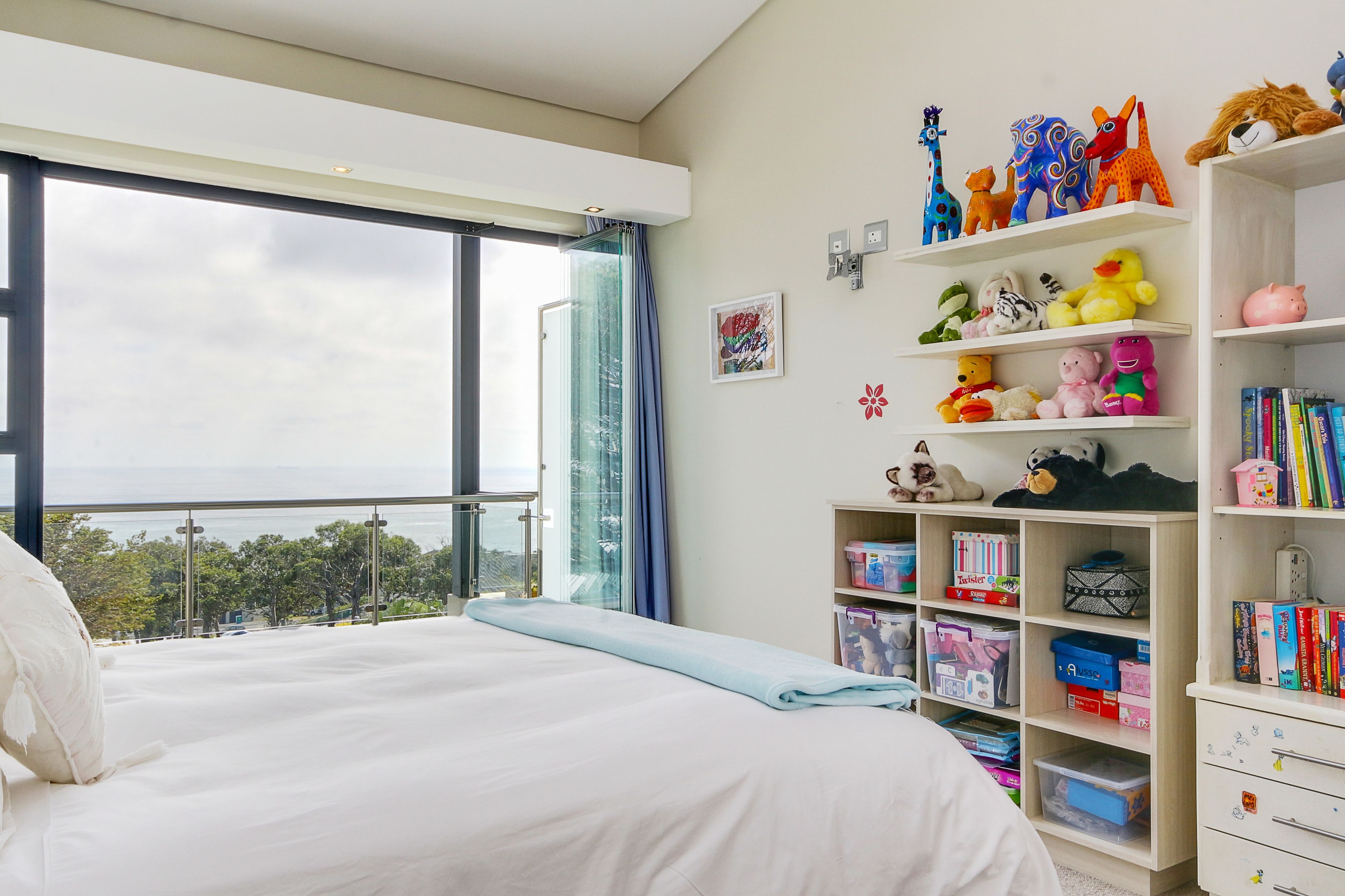A bedroom with a large window, a bed, a bookshelf with toys and books, and a view of the outdoors.