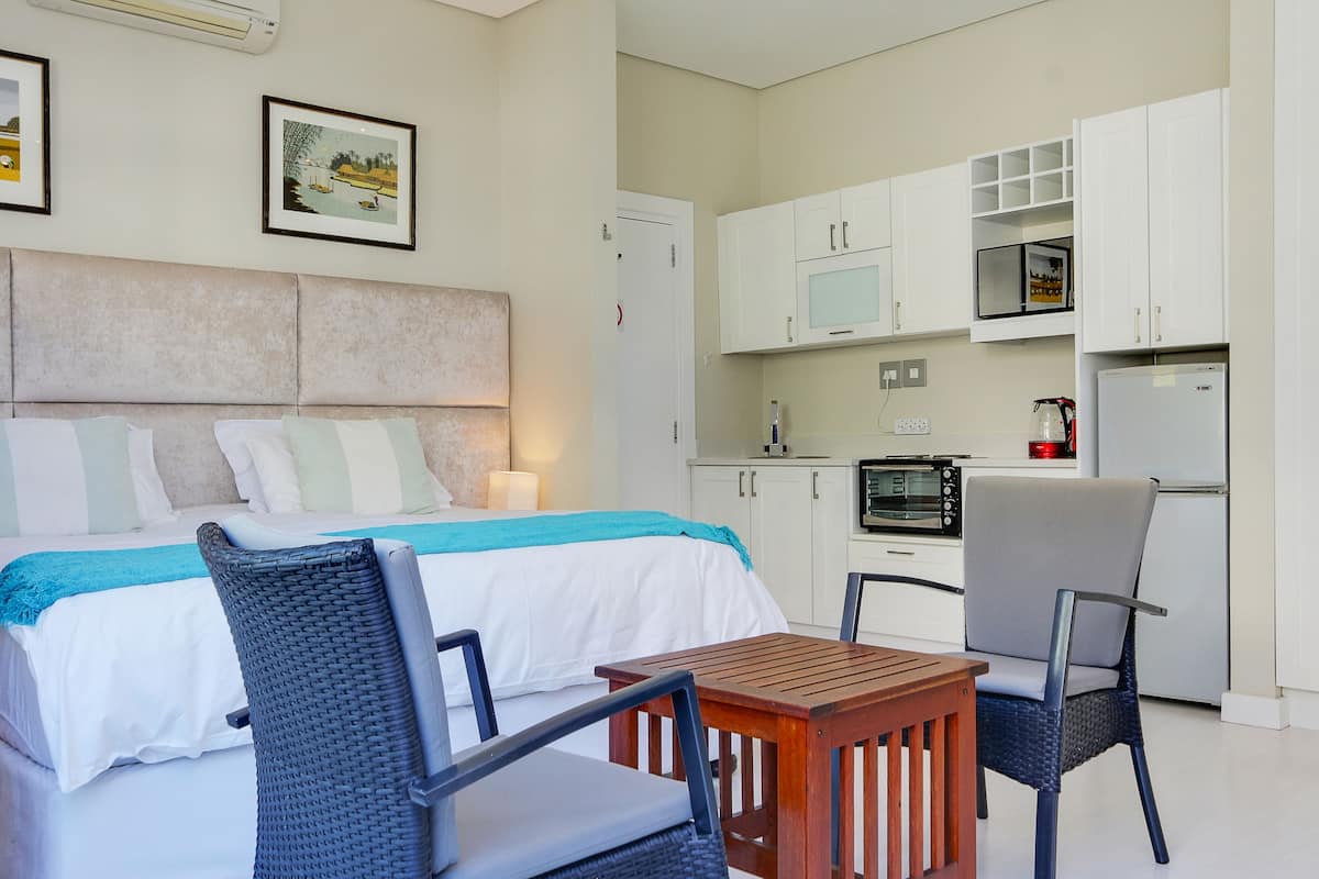 A hotel room with a bed, two chairs, a wooden table, a kitchen area with cabinets, a microwave, and a refrigerator.