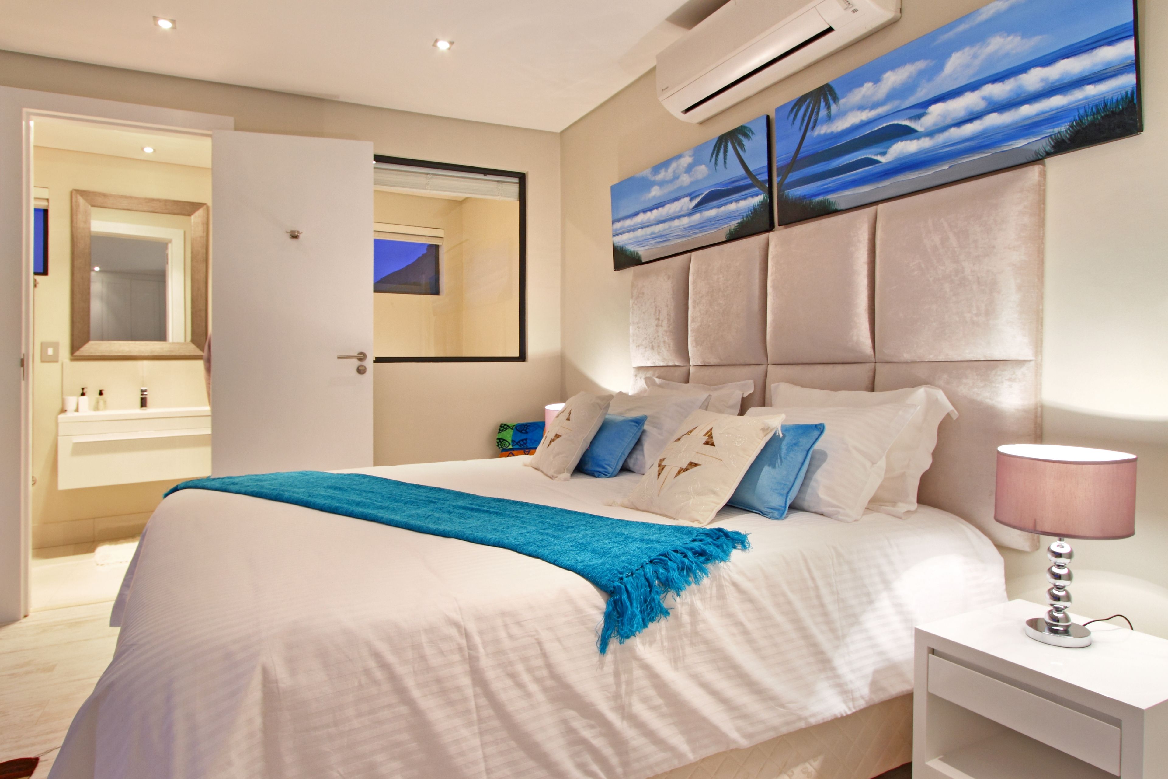 Una camera da letto con un letto, un comodino, una lampada, un quadro raffigurante una spiaggia e una porta aperta che dà su un bagno.