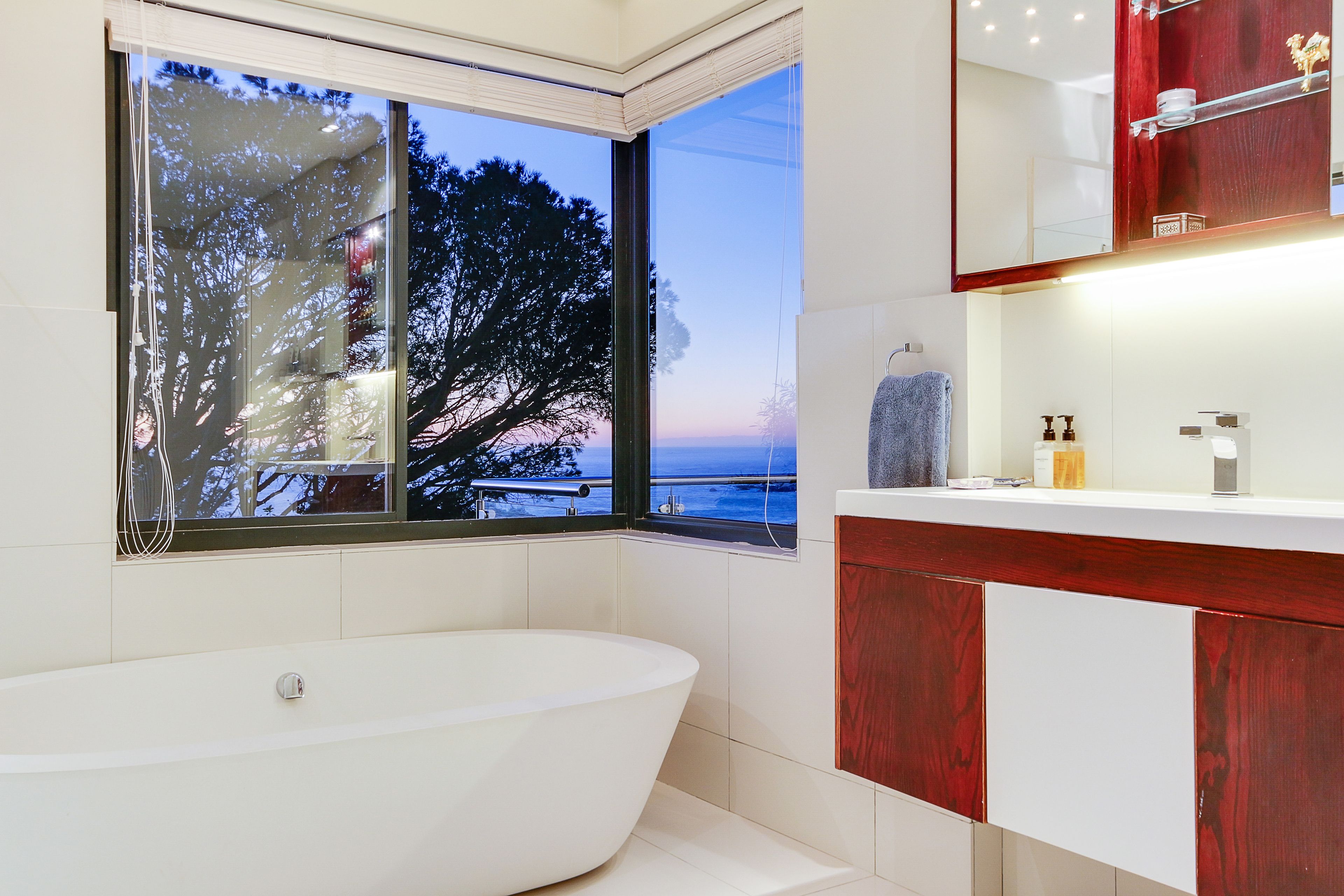 A modern bathroom with a freestanding bathtub, a large window with a view of trees and the sky, and a vanity with a mirror and toiletries.