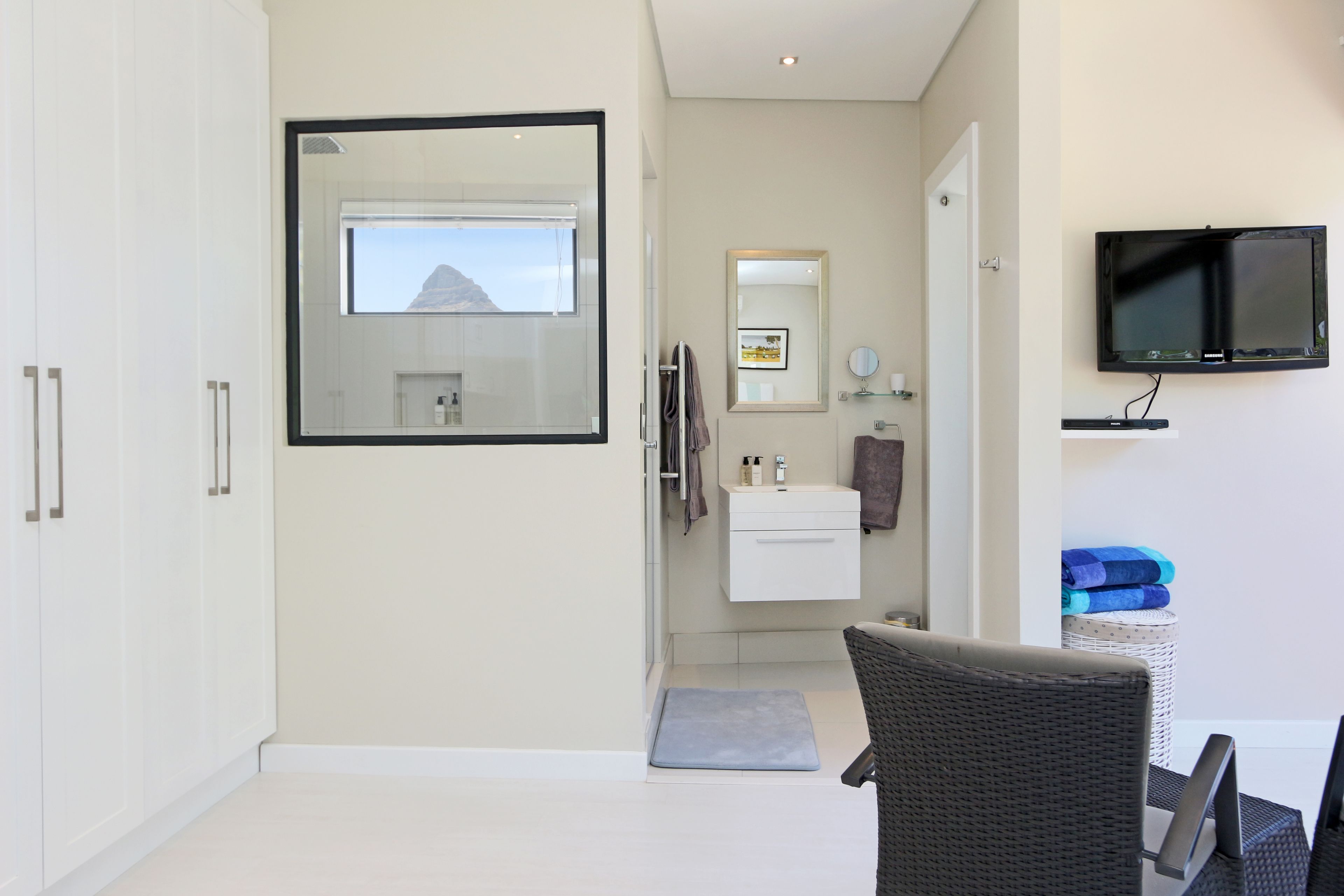 A modern bathroom with a large mirror, a sink, and a toilet. There is a television mounted on the wall and a wicker chair in the room.