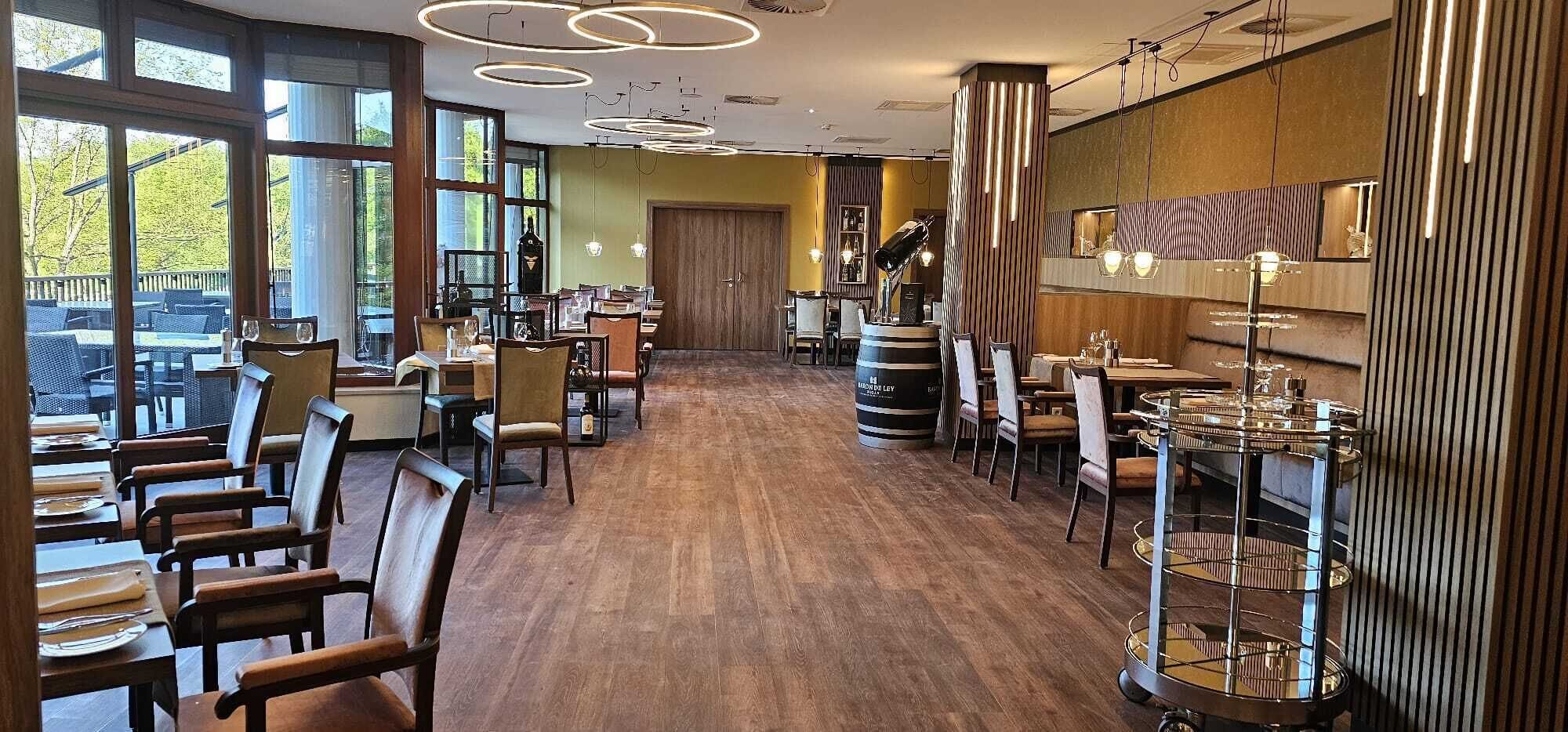 A restaurant with wooden flooring, round tables, and chairs. There are large windows on one side, allowing natural light.