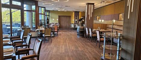 A restaurant with wooden flooring, round tables, and chairs. There are large windows on one side, allowing natural light.