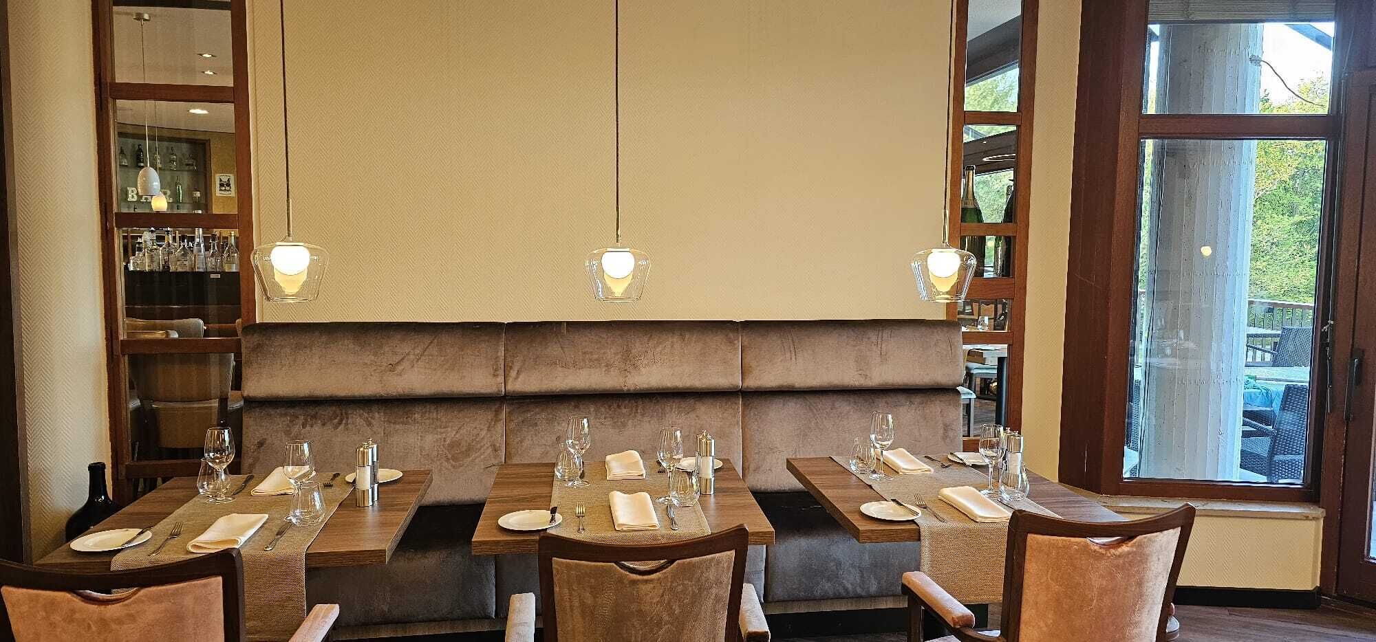 A restaurant with wooden tables and cushioned booths, hanging pendant lights, and a view through large windows.