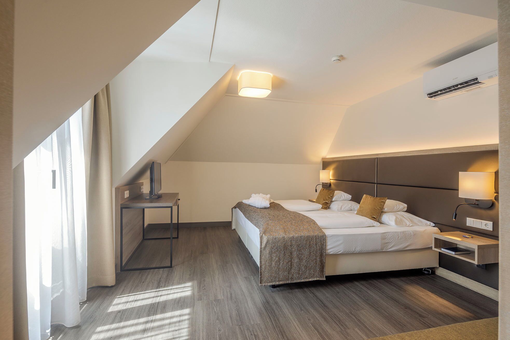 A modern hotel room with a sloped ceiling, a bed with white linens, a bedside table with a lamp, and a TV on a stand.